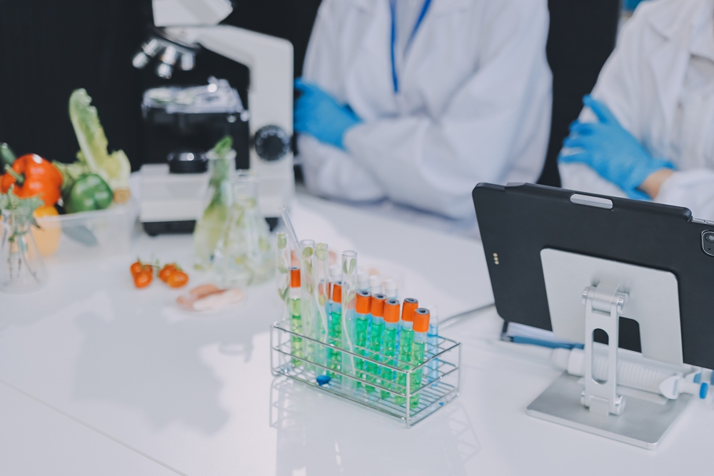 Scientists in lab coats and blue gloves work at a white lab bench with test tubes in a rack and a touchscreen monitor nearby.