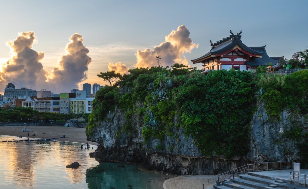 Temple on a green cliff overlooking a sandy beach and calm water with a distant cityscape at sunset