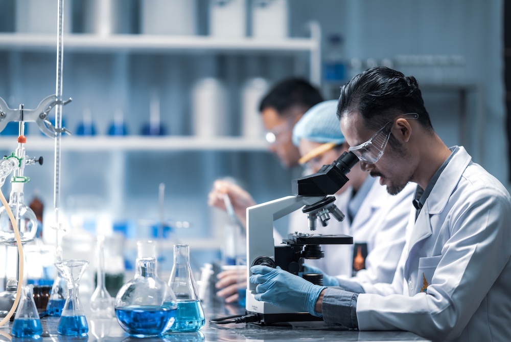 Scientist in safety goggles and a lab coat uses a microscope at a chemistry bench, with blue liquid in glass flasks nearby.
