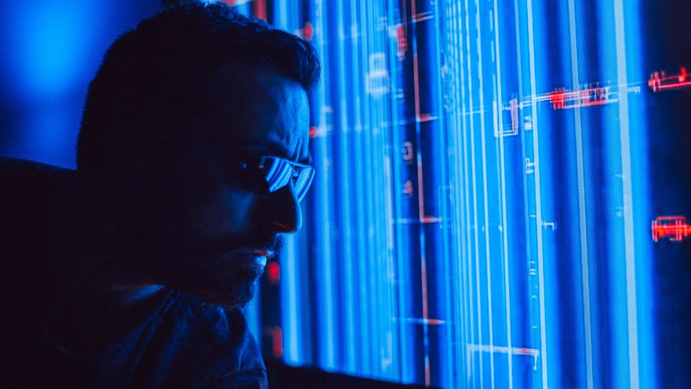 Tech worker in dark clothing and glasses studies a glowing blue data wall or server screen.
